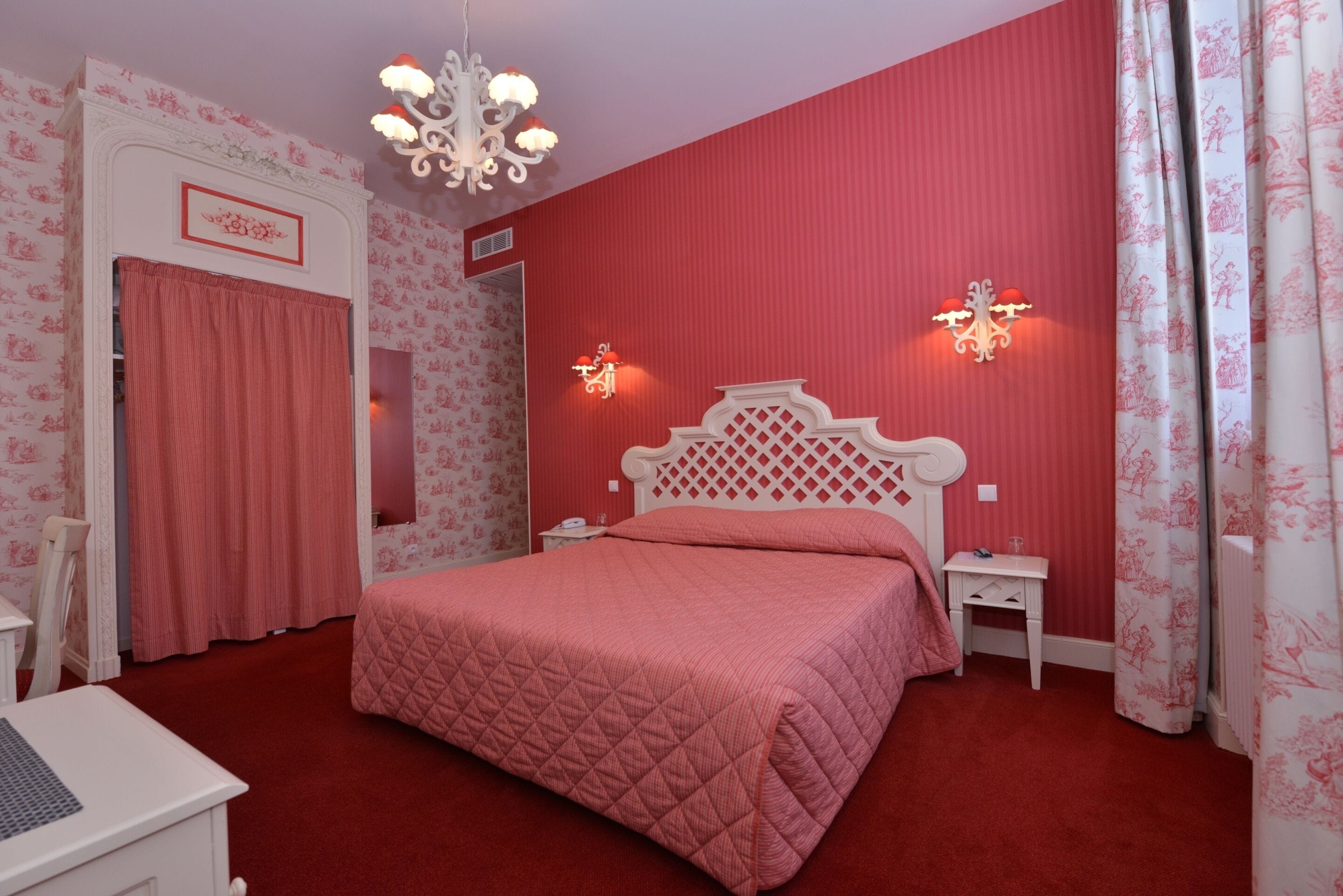 A bedroom with a red wall, a white bed with a diamond-patterned quilt, two bedside tables with lamps, a white dresser, and a chandelier.