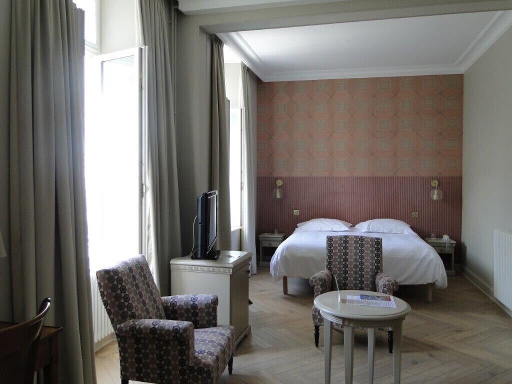 A hotel room with a bed, two chairs, a small table, a TV, and a window with curtains.