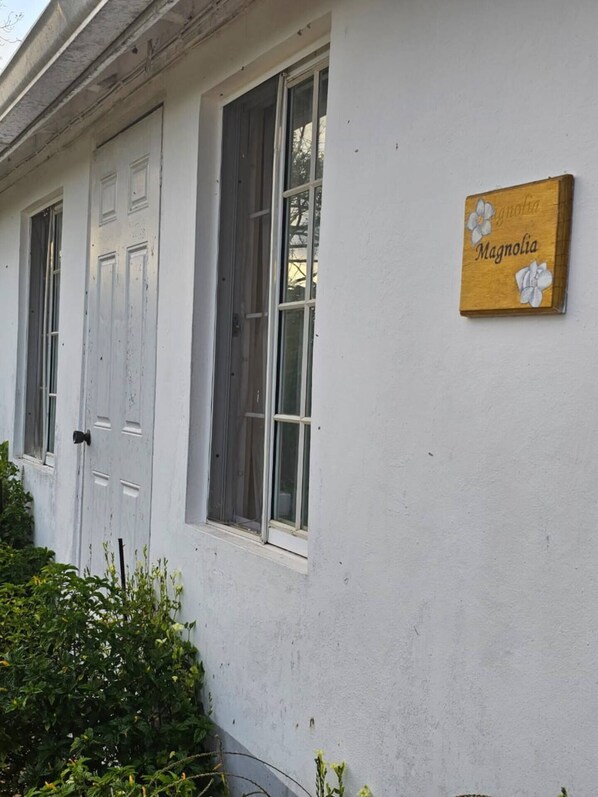 A building with white exterior walls, a wooden sign with the name "Magnolia," and a window with a metal screen. - Dalkeith Cottages-10 Minutes From Mandeville (Knockpatrick)