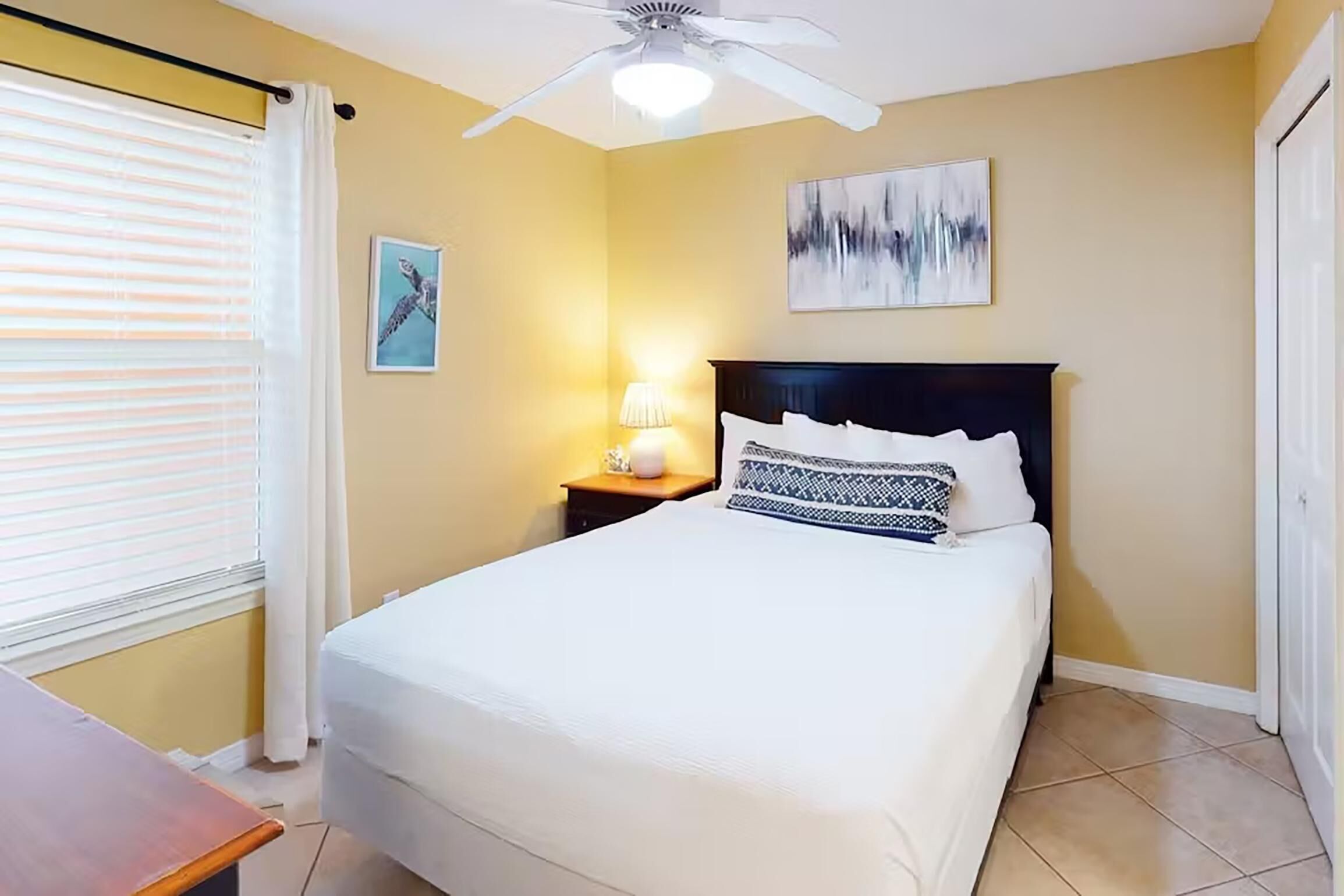A hotel room with a bed, a nightstand, a ceiling fan, a window with blinds, and a wall with two artworks.