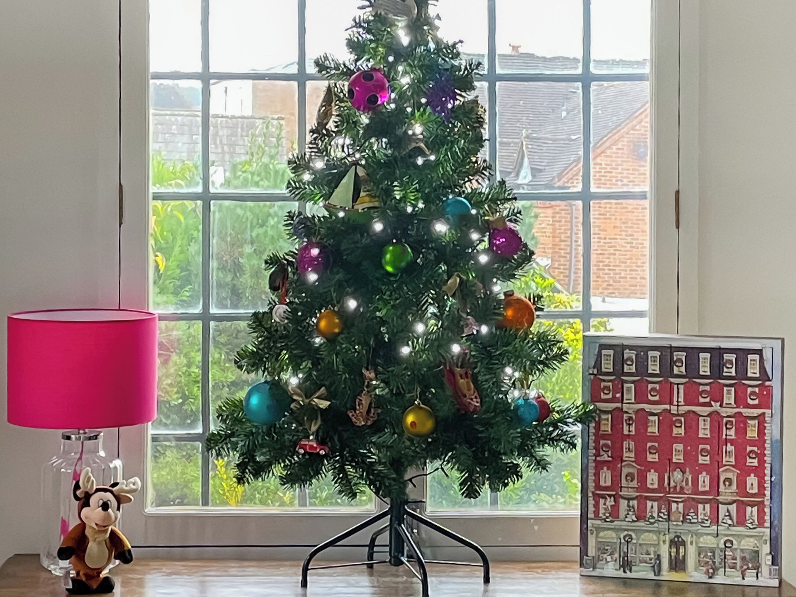 Un árbol de Navidad con luces y adornos, una lámpara rosa y un modelo a escala de un pueblo.