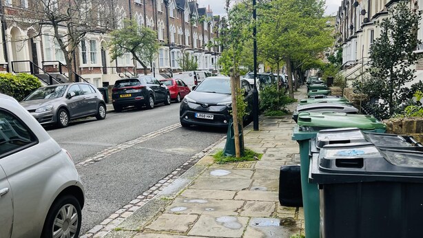 Une rue résidentielle bordée de voitures garées et d’une rangée de poubelles vertes sur le trottoir.
