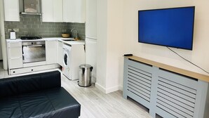Une cuisine compacte équipée d’un four, d’un micro-ondes et d’un lave-linge intégrés, d’un canapé en cuir noir et d’un téléviseur fixé au mur.