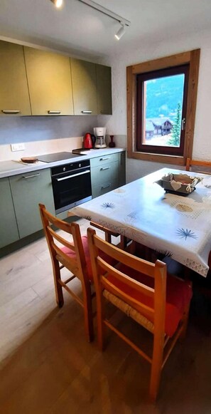 Cocina compacta con armarios verdes, una mesa de comedor de madera y una ventana con vistas a una casa y árboles.