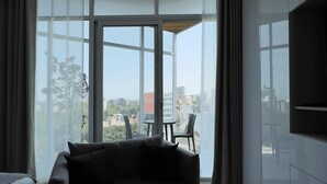 A modern hotel room with a large window, a sofa, a TV, and a dining area with chairs.
