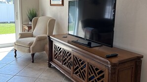 Een woonkamer met een televisie op een houten kast, een beige fauteuil en een potplant.