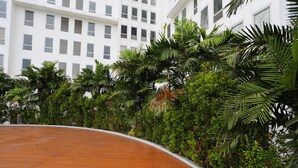 A curved wooden deck with dense tropical foliage and a white building in the background. - Nusantara Rooms By Reccoma (Bekasi)