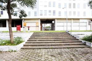 A building with a set of concrete steps leading to a covered entrance, surrounded by a paved area with hexagonal tiles and some greenery.