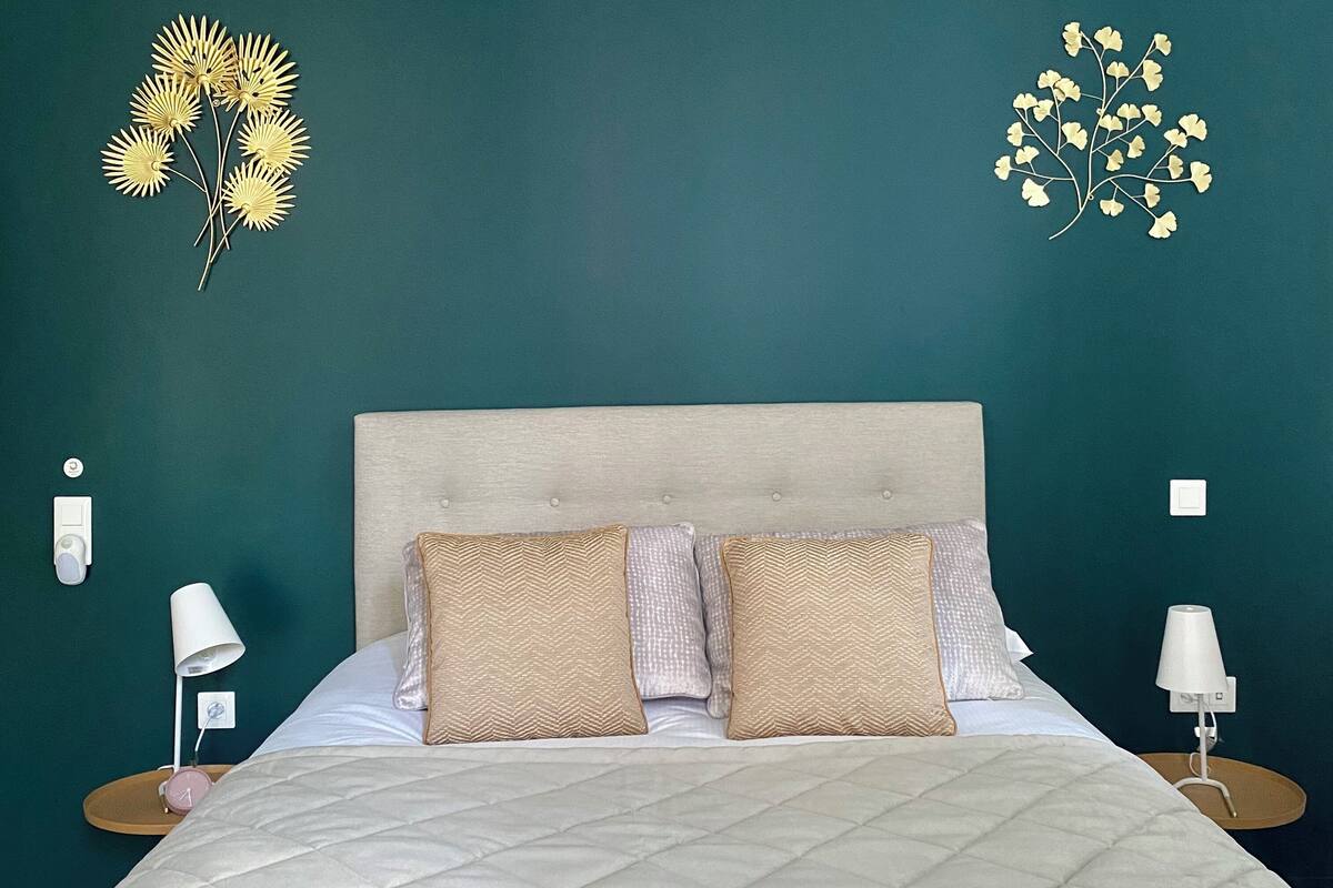 A bed with a quilted duvet, two patterned pillows, and a headboard. There are bedside tables with lamps and a wall clock on the left.