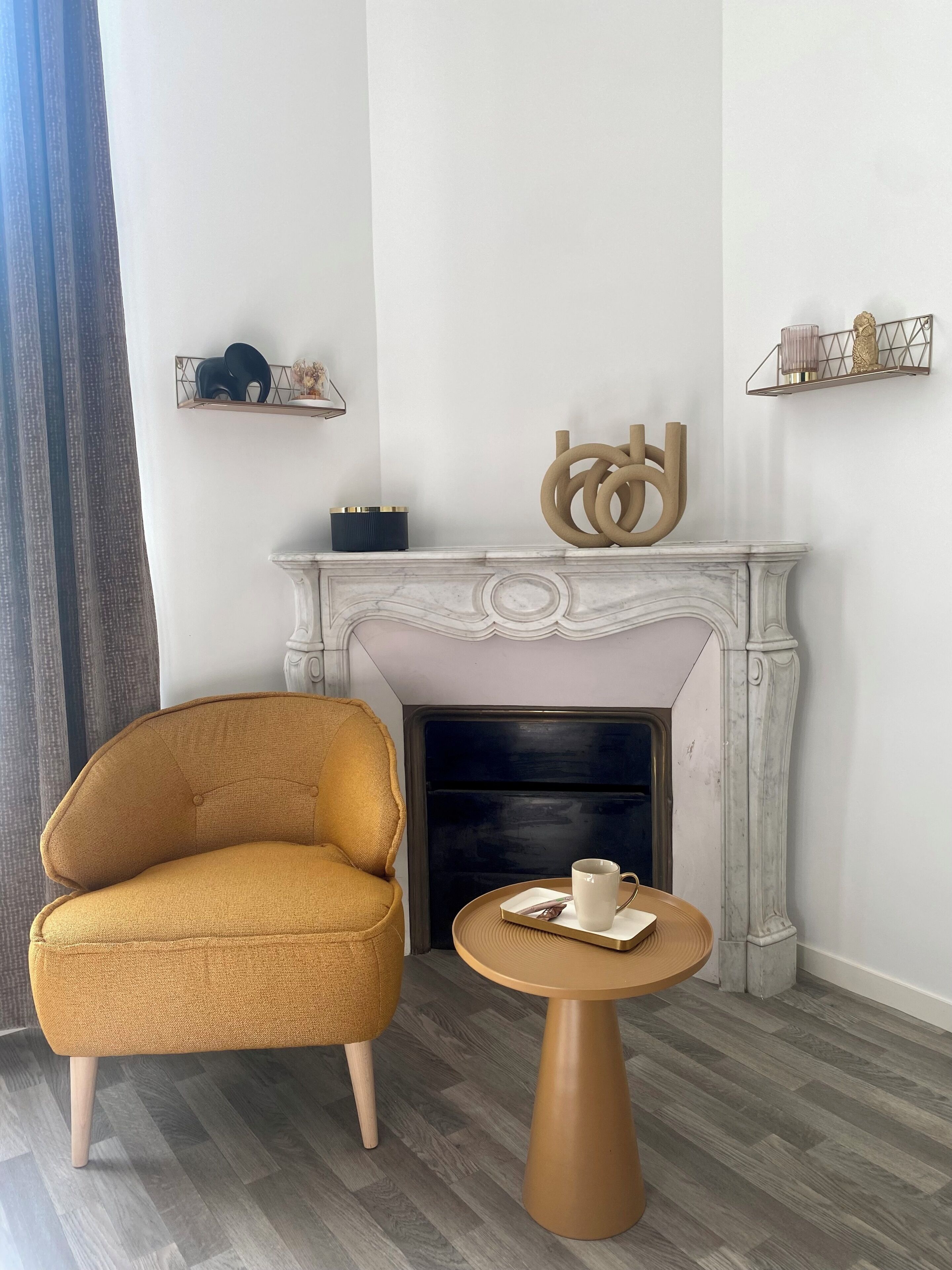 Una habitación con chimenea, un sillón color mostaza y una pequeña mesa redonda con una taza y un platillo.