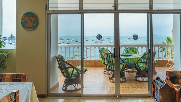 A room with a sliding glass door leading to a balcony with a view of the sea and boats. There are wicker chairs and a table on the balcony. - Apartment - Sleeps & Free Parking (Salinas)