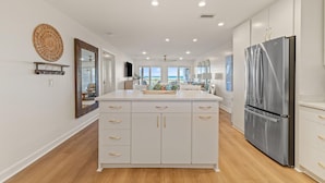 A modern kitchen with white cabinetry, a stainless steel refrigerator, and a large island with gold handles. - Needle Rush Point D11 (Pensacola)