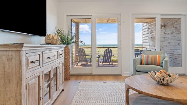 A living room with a large TV, a wooden cabinet, a plant, and a view of the ocean through glass doors. - Needle Rush Point D11 (Pensacola)
