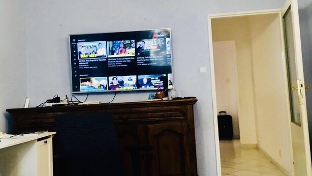 Een kamer met een pronkstuk, een bureau met een computer, een stoel en een televisietoestel waarop verschillende zenders te zien zijn.