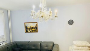 Een woonkamer met een zwarte leren bank, een glazen salontafel, een wit bed met kussens, een kroonluchter, een schilderij en een klok aan de muur.