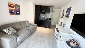 A modern living room with a grey sofa, a flat-screen TV, and a kitchen in the background.