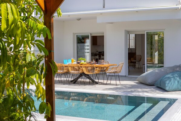 A modern outdoor pool area with a dining table and chairs, surrounded by glass doors leading to a kitchen and a living area.
