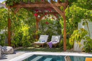A wooden pergola with two white cushioned chairs under lush green foliage.