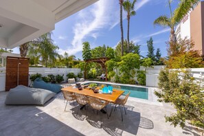 A patio with a wooden table and wicker chairs, a pool, and lush greenery.