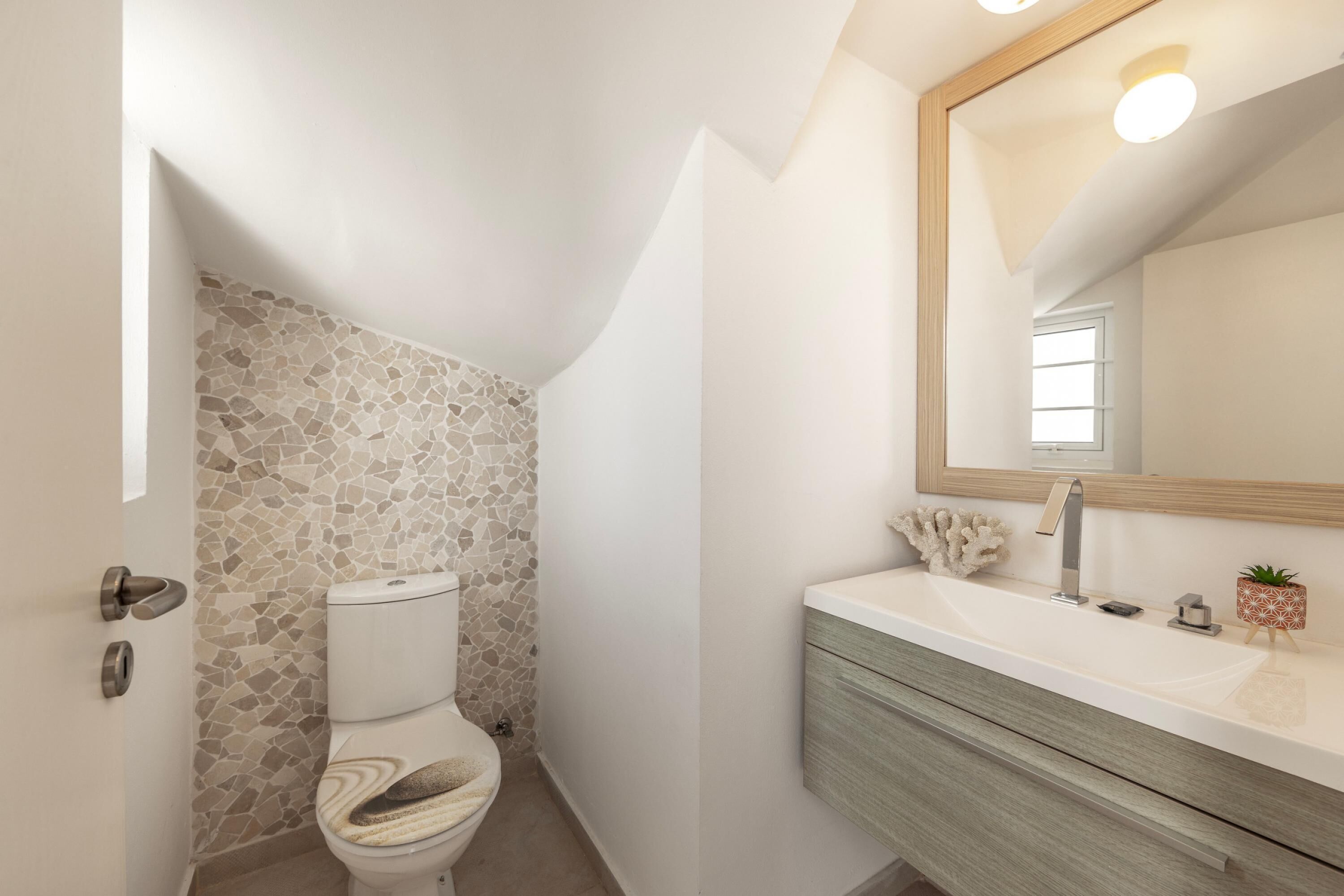 A bathroom with a stone mosaic wall, a white toilet, a wooden vanity, and a mirror with a light fixture.