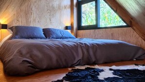 A cozy attic bedroom with a wooden sloped ceiling, a large window, a bed with a dark-colored comforter, and a fur rug on the floor. - Cabañas Grasshopper SHIELD (Tepoztlán)