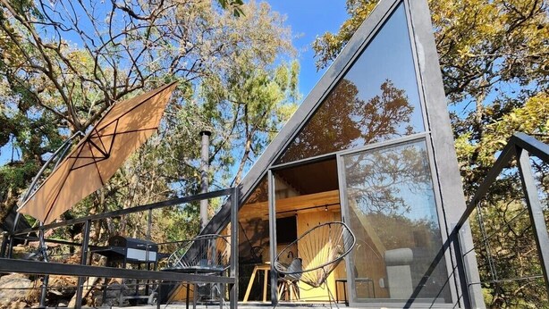 A modern, glass-walled cabin with a metal frame, surrounded by trees. - Cabañas Grasshopper SHIELD (Tepoztlán)