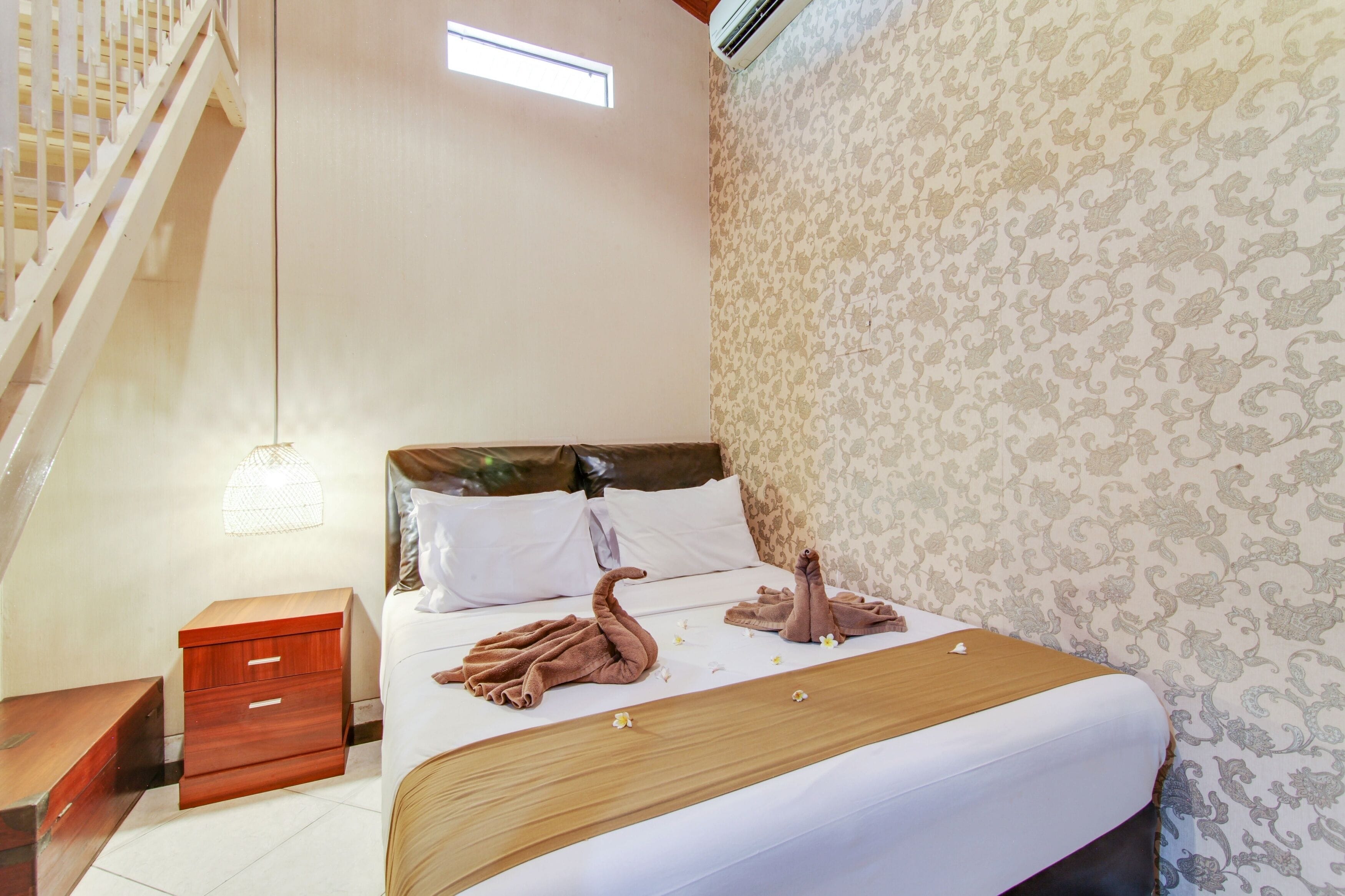 A hotel room with a bed, a nightstand, a wooden dresser, a staircase, and patterned wallpaper.