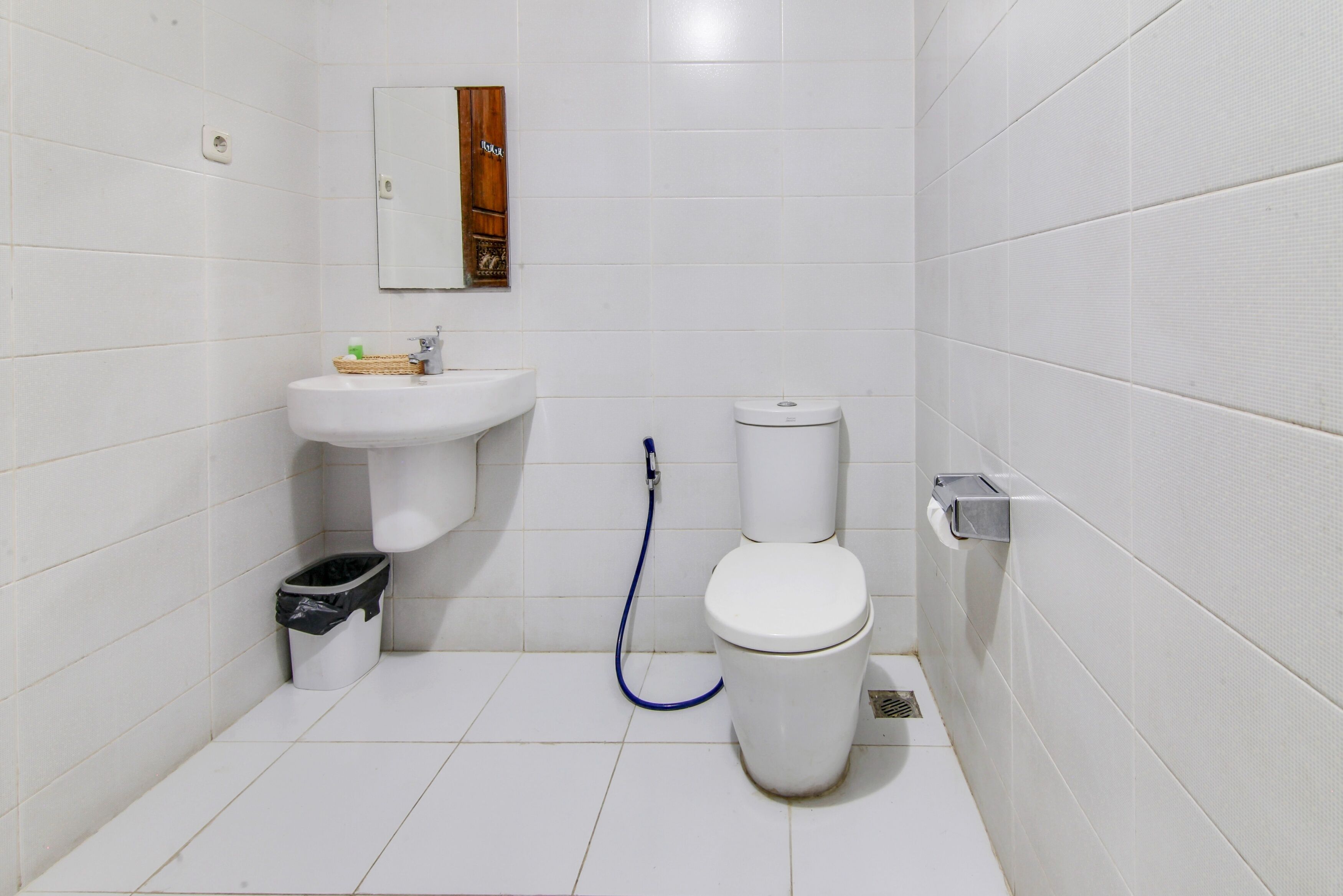 A small, clean bathroom with white tiles, a sink, a toilet, and a trash bin.