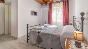 A bedroom with a bed, a bedside table with a lamp, a TV on the wall, and a window with curtains. - Villa Ariana - Standard Room (Gatto Corvino)