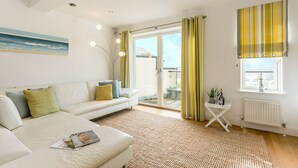 A modern living room with a white sofa, a small side table, a large window with curtains, and a painting on the wall. - Beachside luxury apartment with sea view balcony and parking (Padstow)