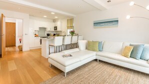 A modern living room with a white sofa, a small dining table, and a kitchen in the background. - Beachside luxury apartment with sea view balcony and parking (Padstow)