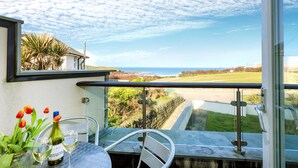 A balcony with a view of the sea, a table with wine glasses and tulips, and a metal chair. - Beachside luxury apartment with sea view balcony and parking (Padstow)