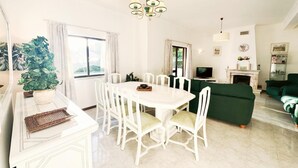 A bright living room with a white dining table and chairs, a green sofa, a fireplace, and a television. - Villa Cavaco in Quarteira (Loulé)