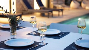 Une table à manger dressée avec de l’argenterie blanche, des verres à vin et des couverts, donnant sur un espace piscine agrémenté de chaises longues.