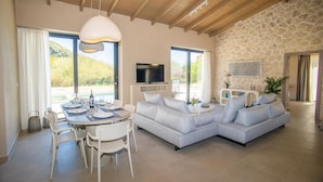 Un salon moderne avec un mur en pierre, un canapé d’angle, une table à manger avec des chaises et un téléviseur fixé au mur.