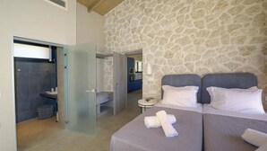 Une chambre d’hôtel moderne avec un mur en pierre, un grand lit avec une literie grise, une petite table ronde et une salle de bain séparée dotée d’une porte en verre.