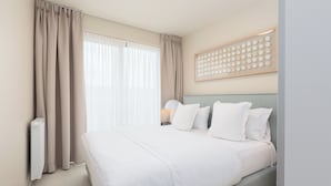 A neatly made bed with white linens, a headboard with a circular pattern, and a window with sheer curtains. - Casselbergh Residences Sky Bridge (Ostend)