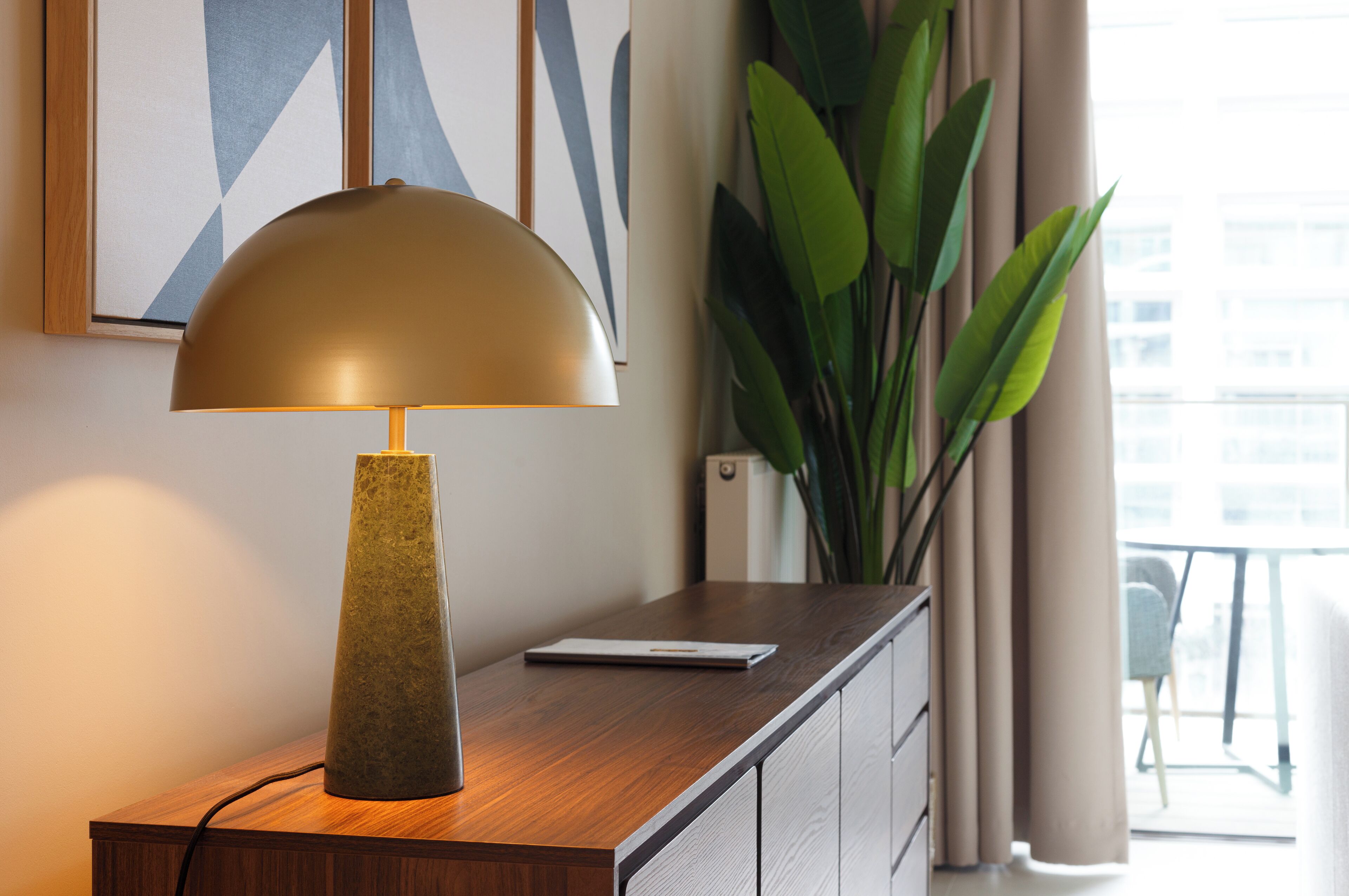 Una lámpara de escritorio con una base texturizada y una pantalla en forma de cúpula, sobre una superficie de madera junto a un dispositivo blanco.
