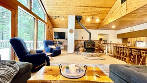 A cozy living room with a fireplace, wooden ceiling, and large windows overlooking a deck. - “Big Trees Retreat,” Boasts 5BR/2.5BA, Sleeps-16. 2-level living. Dog friendly! (Camp Connell)