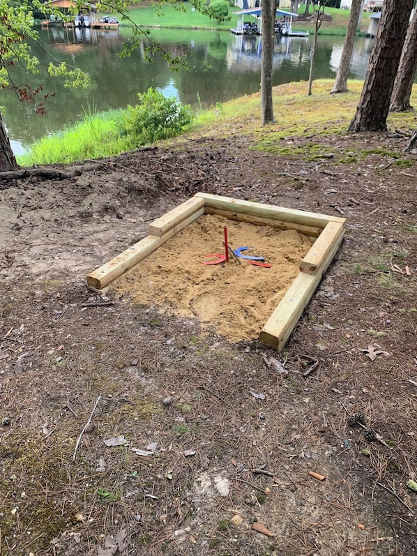 A sandbox with wooden borders and a shovel, surrounded by natural ground and trees.