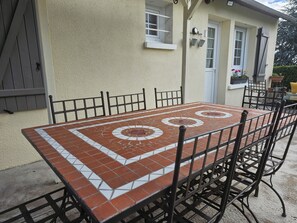 Une terrasse avec une table et des chaises carrelées, un petit jardin avec des plantes en pot et une maison à l’extérieur beige et aux bordures blanches.