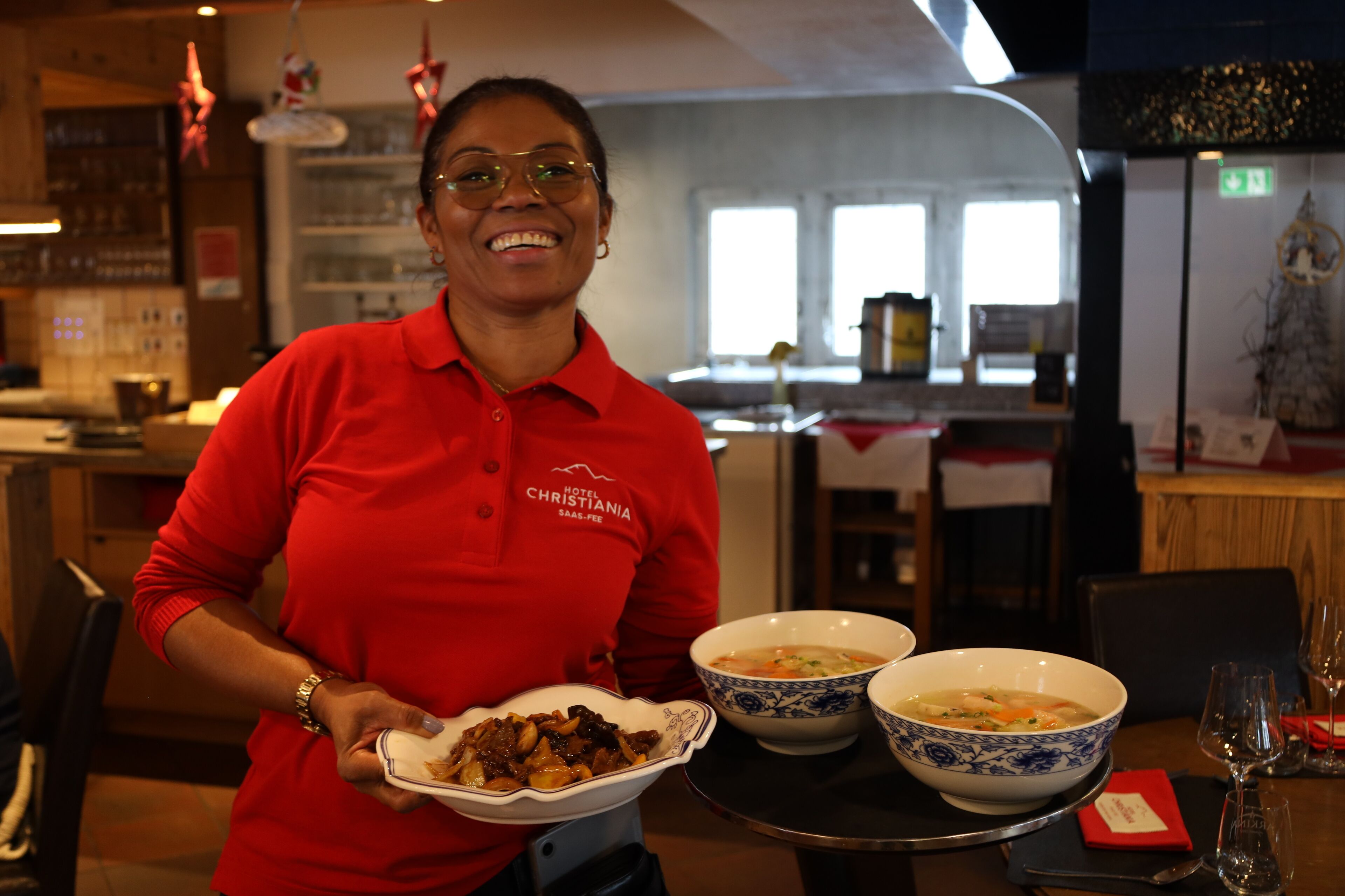 A person in a red shirt holding a plate with food and two bowls of soup on a table.