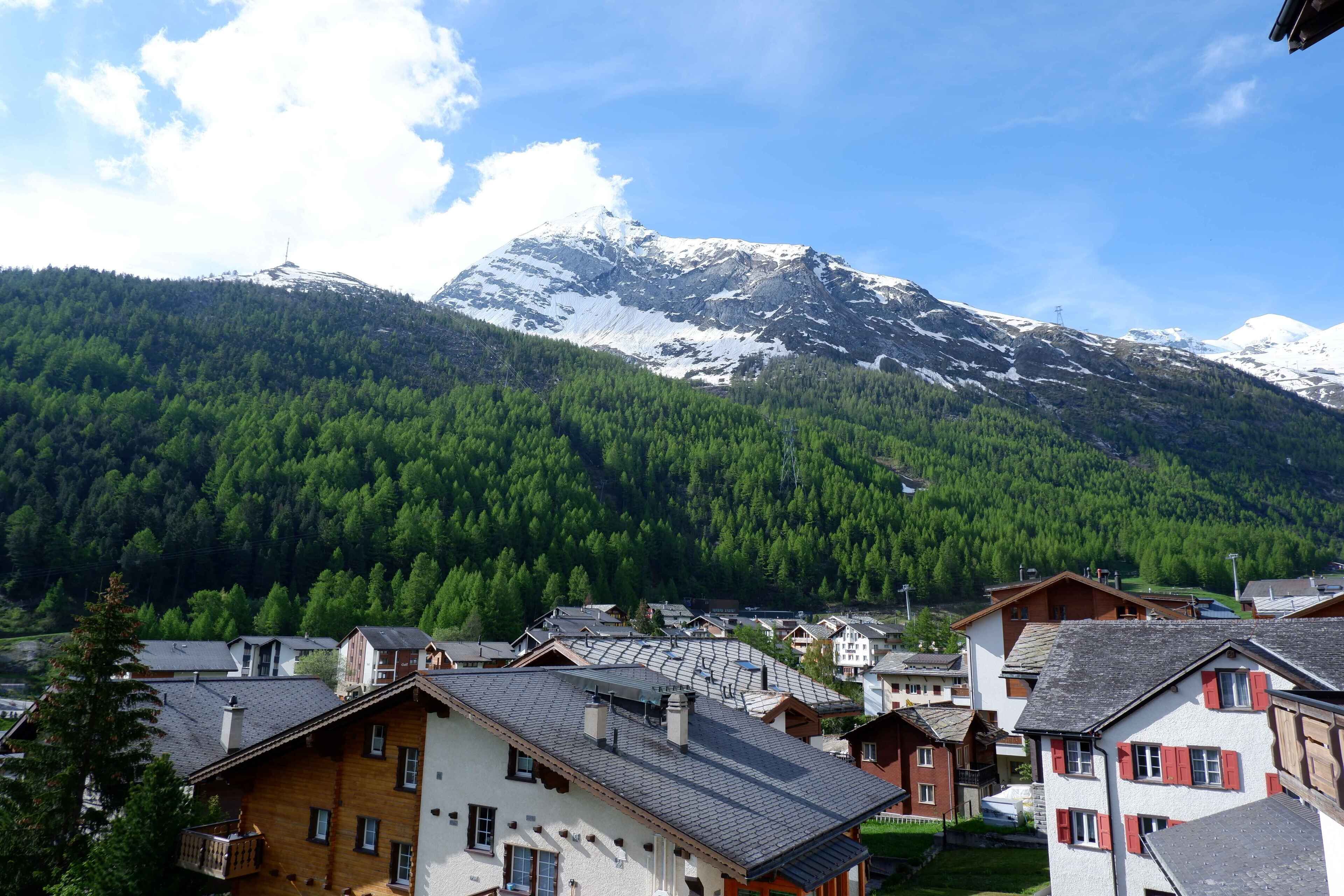 A mountain village with traditional wooden houses, a clear sky, and a snow-capped mountain in the background.
