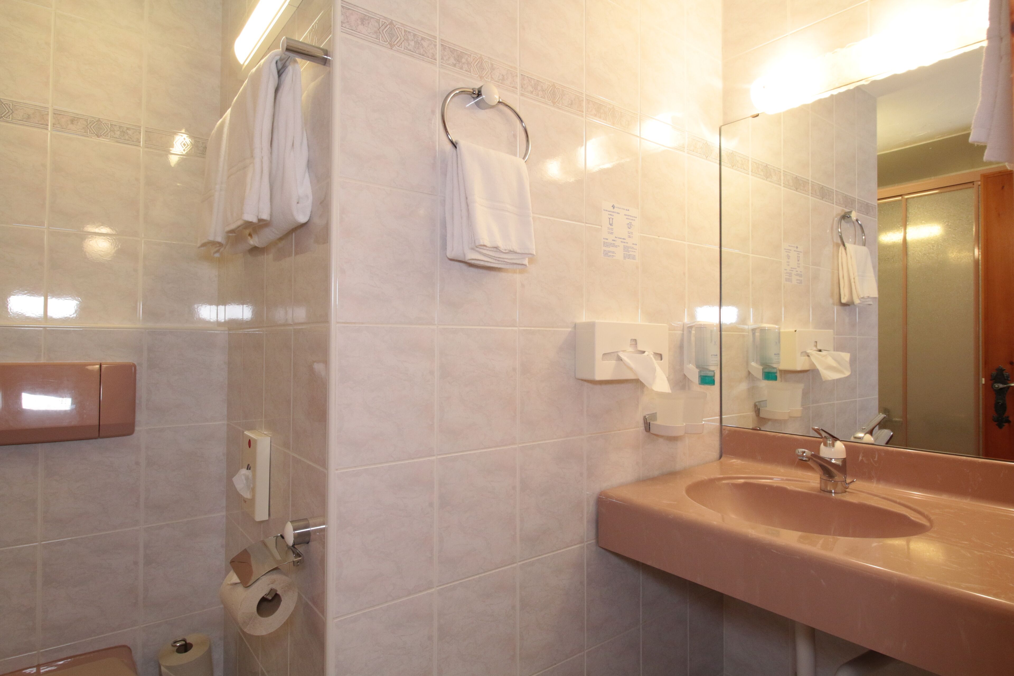 Un baño con lavamanos, espejo, toallero y dispensador de papel higiénico.