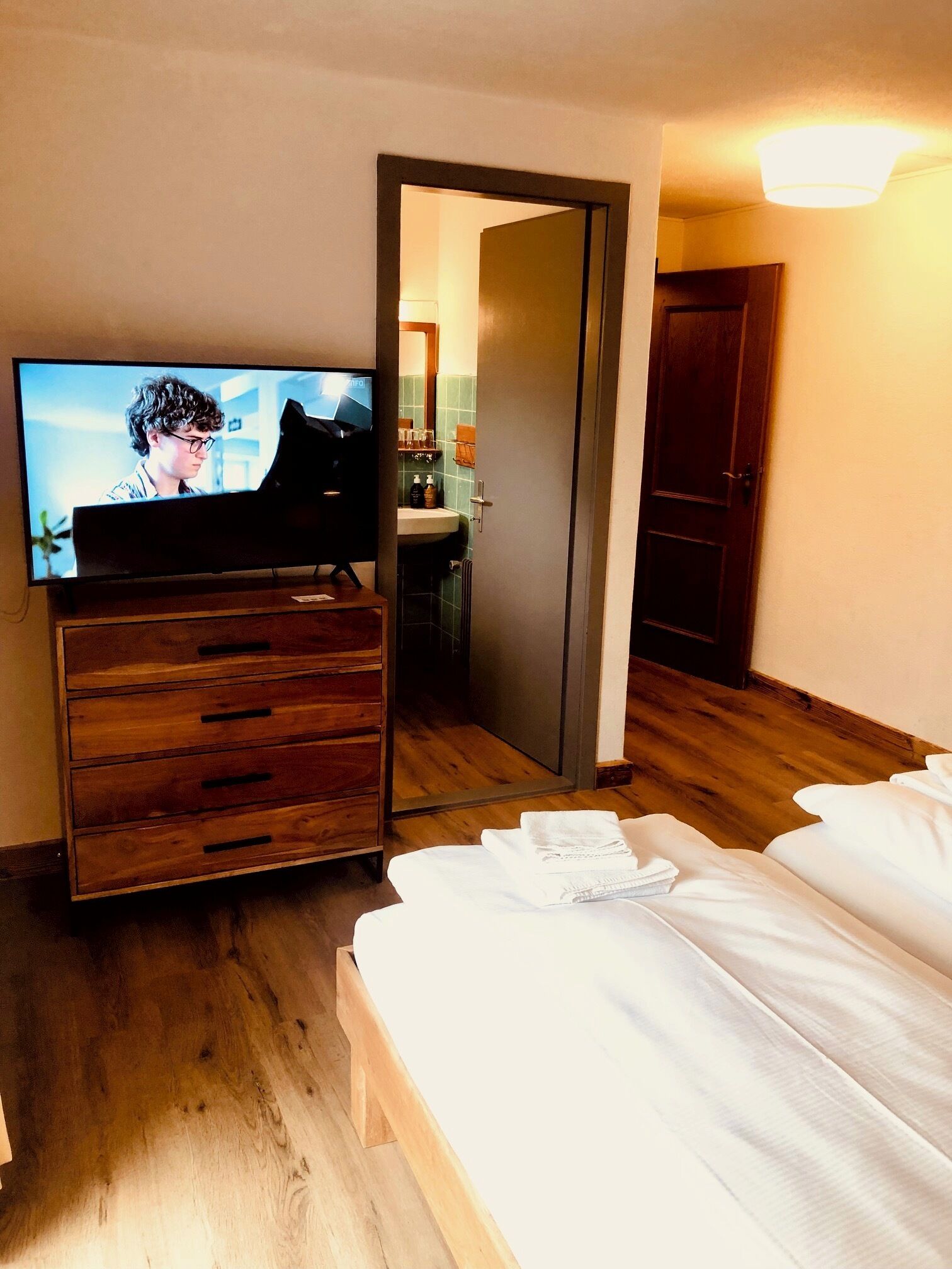 A hotel room with a wooden dresser, a television, a bed with white linens, and a bathroom visible through an open door.