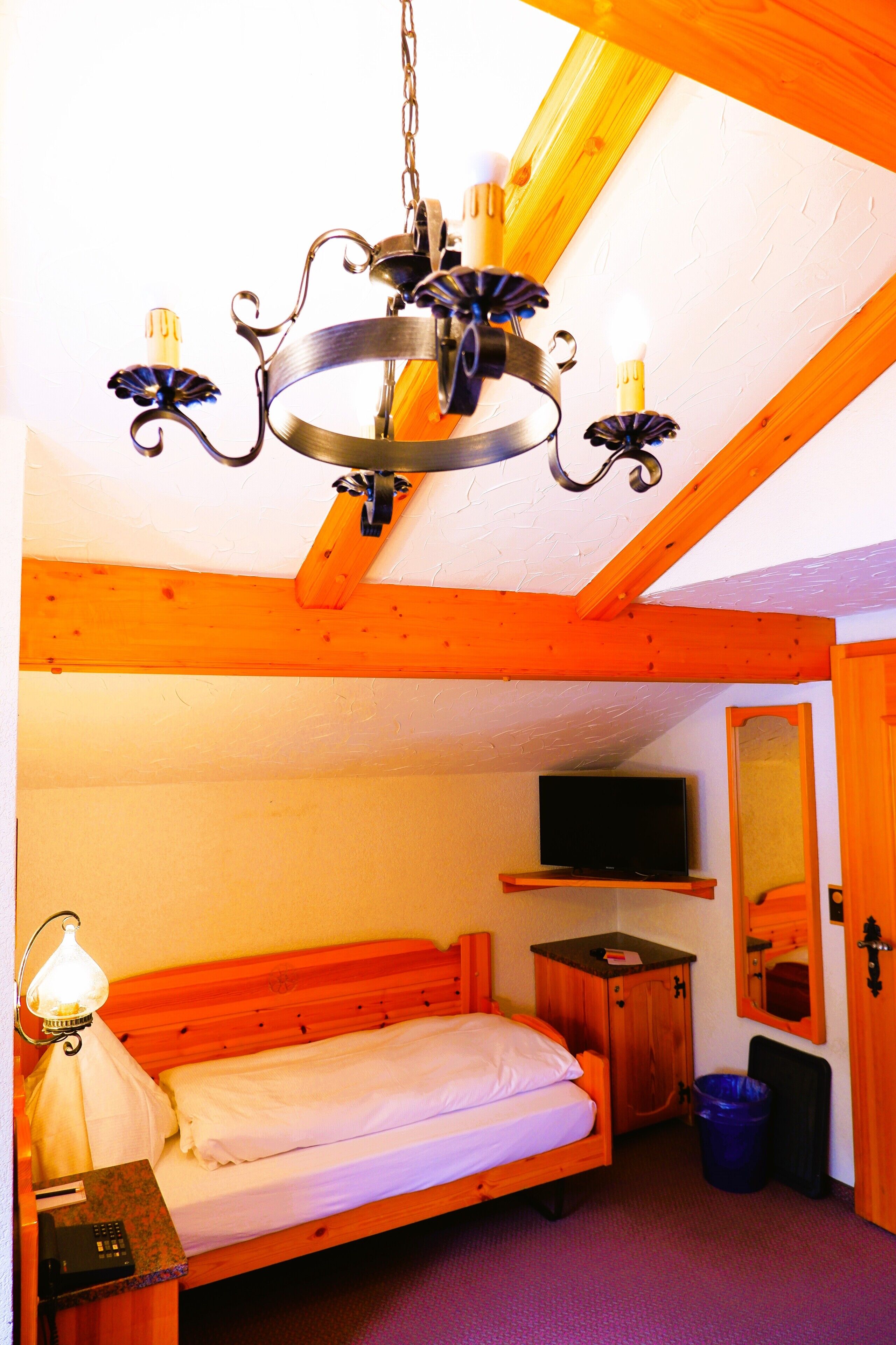 A bedroom with a bed, a television, a wooden cabinet, a trash bin, and a chandelier.