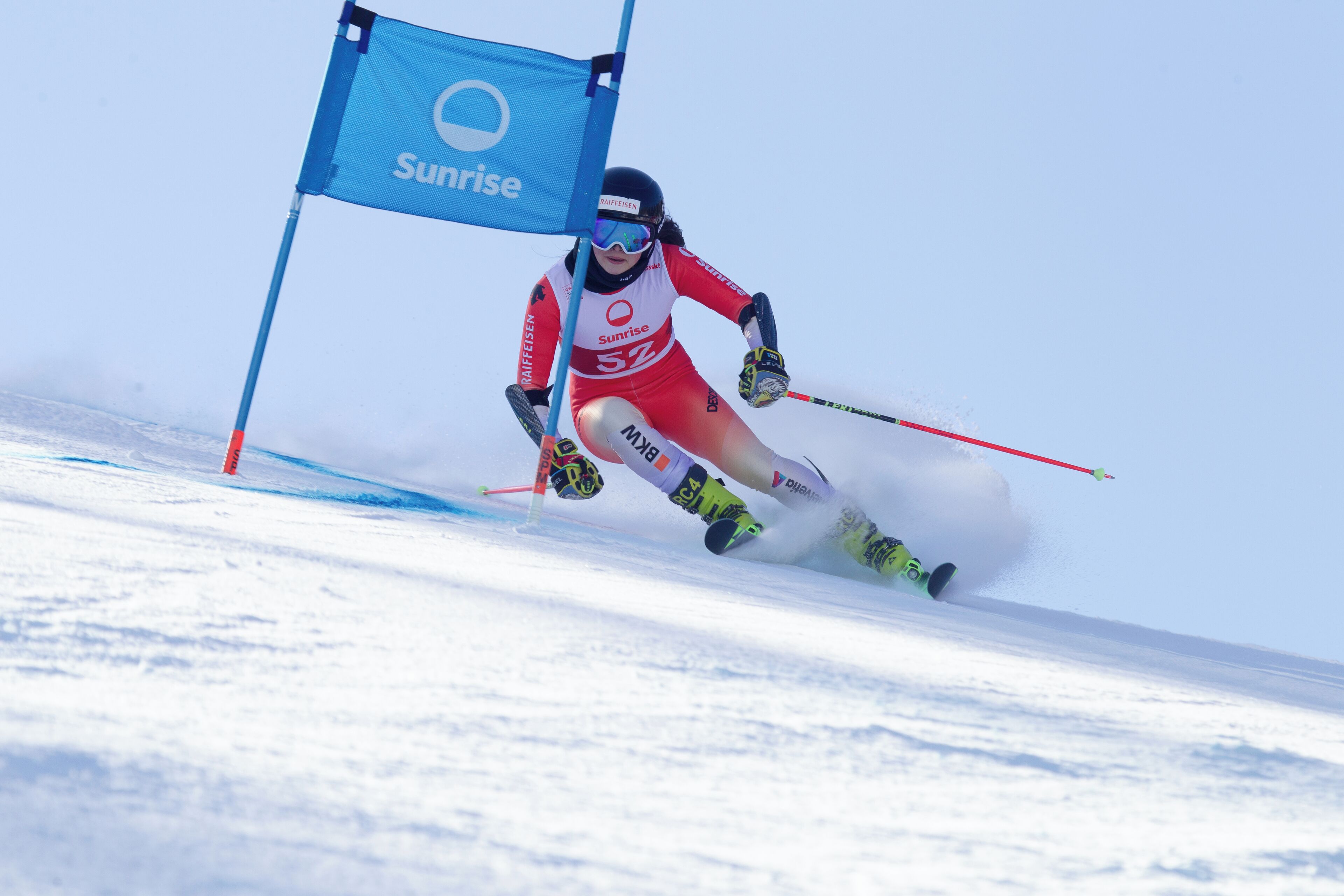 A skier in motion, wearing a red and white uniform, skiing down a snowy slope with a blue flag marked "Sunrise" in the background.
