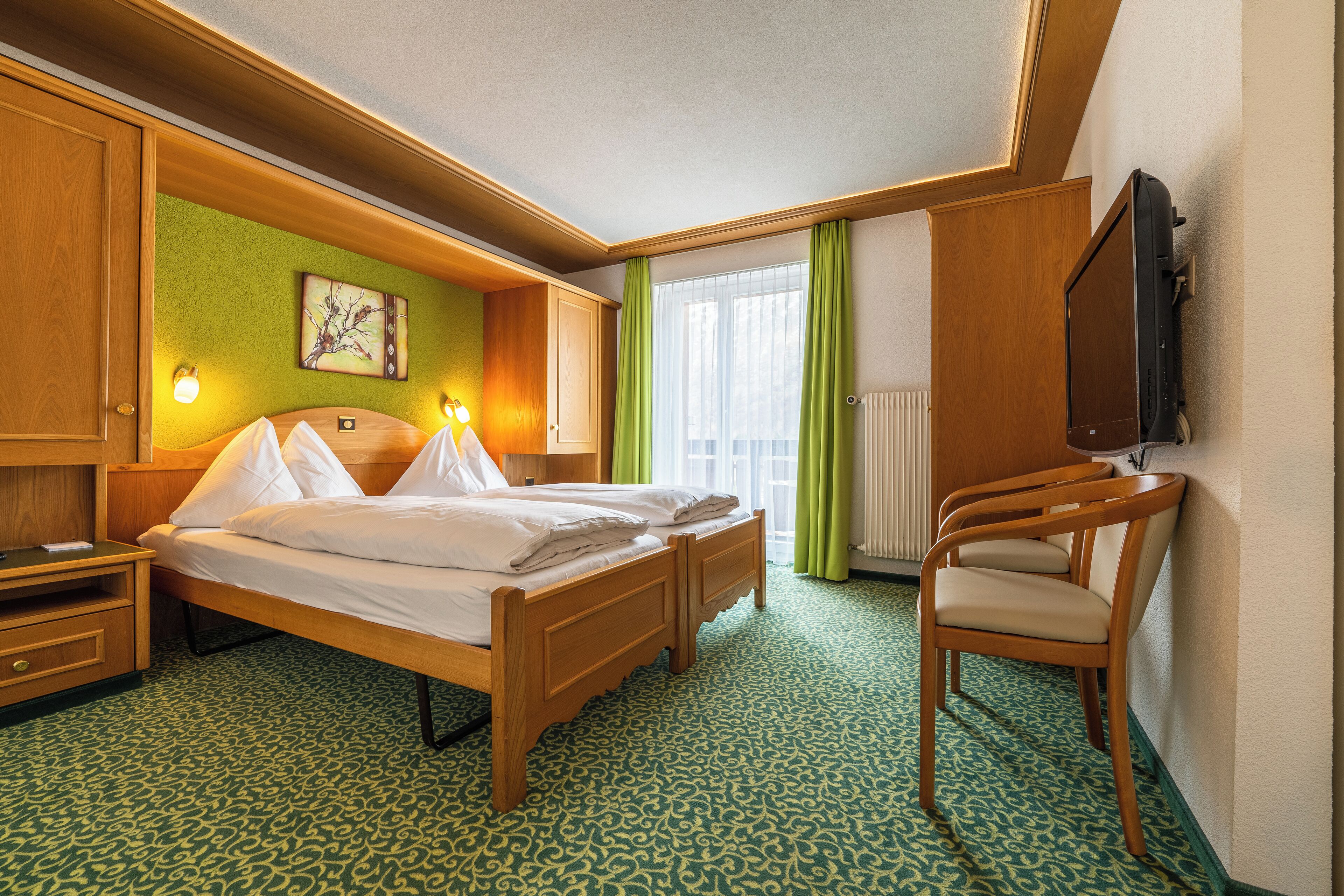 A hotel room with a wooden bed, two bedside tables, a chair, a television, and a window with curtains.