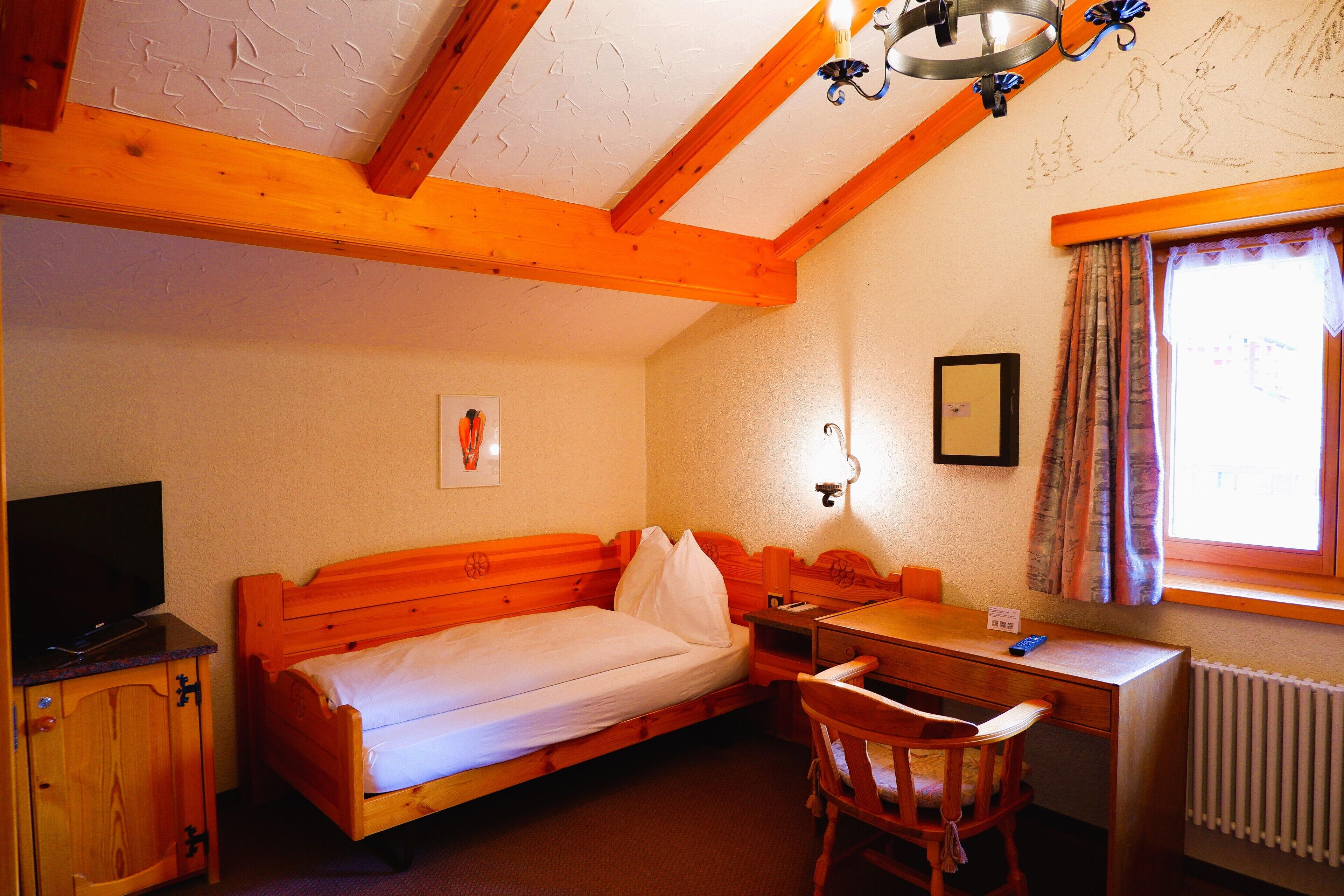 A room with a wooden bed, a desk, a chair, a television, and a window with curtains.