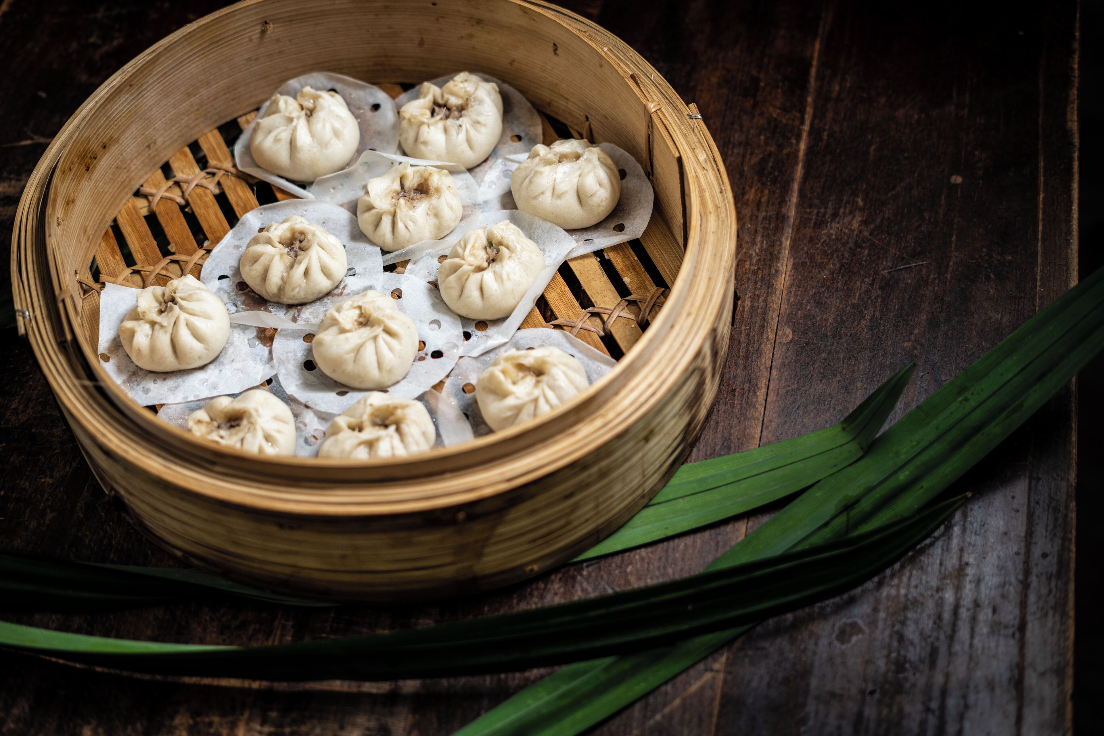 A bamboo steamer with several dumplings on parchment paper, placed on a wooden surface with green leaves beside it.